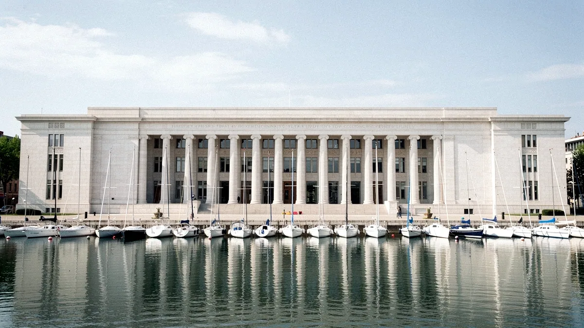 Illustration: A grand pale granite facade with tall columns facing a calm harbour with moored sailboats.