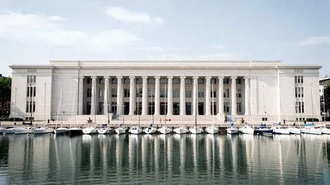 Illustration: A grand pale granite facade with tall columns facing a calm harbour with moored sailboats.
