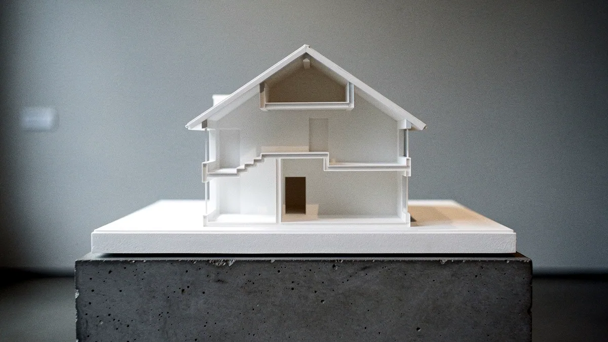 Illustration: A white plaster house cross section cast with stepped room volumes on a concrete plinth.
